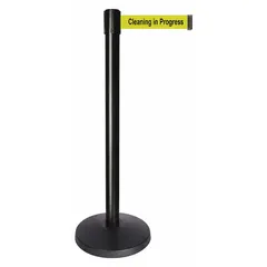 This retractable belt post barrier features a black metal alloy post and base with a 10 ft yellow ABS belt printed with 'Cleaning in Progress' message. The belt retracts into the post when not in use. Used to cordon off areas and communicate temporary access restrictions during cleaning operations.